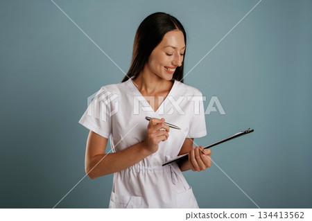Notepad and pen in hands, nurse is standing. Young woman is in the studio against background Notepad and pen in hands, nurse is standing. Young woman is in the studio against background 134413562
