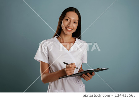 Notepad and pen in hands, nurse is standing. Young woman is in the studio against background Notepad and pen in hands, nurse is standing. Young woman is in the studio against background 134413563