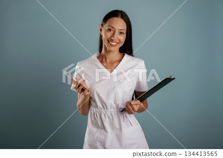 In professional lab coat, nurse is standing. Young woman is in the studio against background In professional lab coat, nurse is standing. Young woman is in the studio against background 134413565