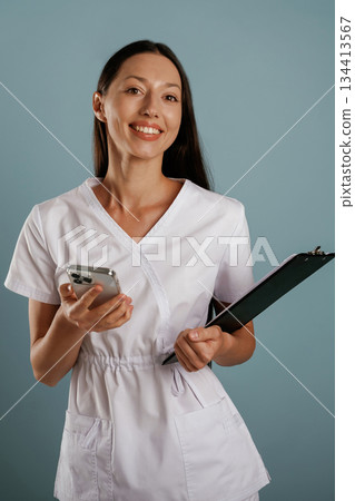 In professional lab coat, nurse is standing. Young woman is in the studio against background 134413567