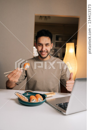 Friendly young man happily giving thumbs up, enjoying delicious sushi dinner while working or relaxing with laptop at home, embracing remote lifestyle and convenience, looking at camera. Friendly young man happily giving thumbs up, enjoying delicious sushi dinner while working or relaxing with laptop at home, embracing remote lifestyle and convenience, looking at camera. 134413573