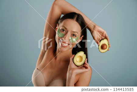 Green patches under eyes, holding avocado. Young woman is in the studio against background 134413585