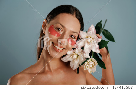 Cleaning patches under eyes, holding lily flowers. Young woman is in the studio against background Cleaning patches under eyes, holding lily flowers. Young woman is in the studio against background 134413598