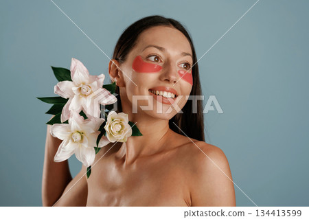 Cleaning patches under eyes, holding lily flowers. Young woman is in the studio against background Cleaning patches under eyes, holding lily flowers. Young woman is in the studio against background 134413599