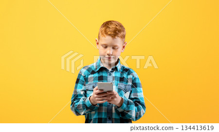 Cute ginger kid with mobile phone on yellow studio background, using exciting mobile applications or chatting with friends, copy space. Happy redhead boy using smartphone, gadget addiction 134413619