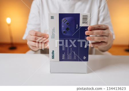 Ufa, Russia - 30 September 2025. Woman hands unboxing new wireless gamepad on white table, revealing controller in package. Concept of modern gaming tech, leisure, excitement of recent purchase. 134413623