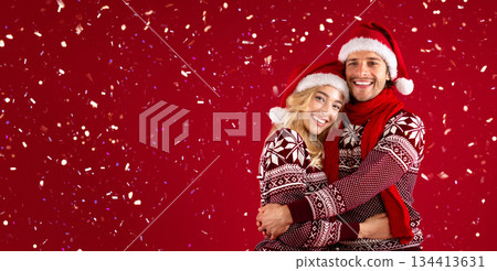 It's Christmas. Beautiful romantic couple in Santa hats and warm sweaters hugging on red background 134413631
