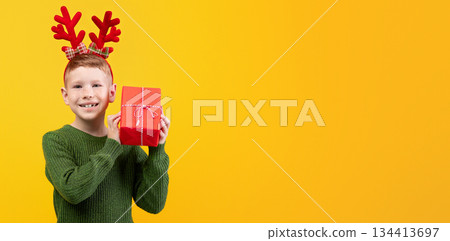 Happy kid with New Year gift over yellow studio background, copy space. Excited boy with deer accessory holding xmas present next to ear, wondering what is inside. Winter holidays celebration 134413697