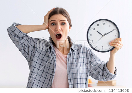 Shocked young lady with clock grabbing her head in terror, standing in messy room after party, too late to clean apartment before return of her parents. Space for text 134413698