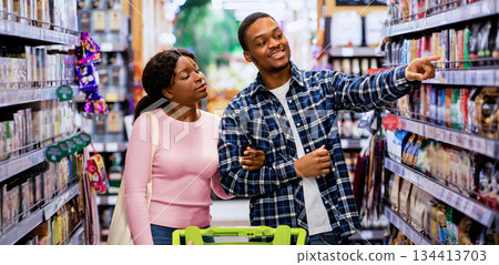 Pretty black woman and her boyfriend shopping for groceries together, selecting goods, pointing at shelves in supermarket. Loving African American couple choosing food at produce section of big mall Pretty black woman and her boyfriend shopping for groceries together, selecting goods, pointing at shelves in supermarket. Loving African American couple choosing food at produce section of big mall 134413703