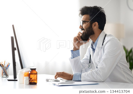 Handsome millennial Arab doctor wearing medical coat working at clinic, general practitioner sitting at workdesk, using computer pc, have phone conversation with patient or colleague, side view 134413704