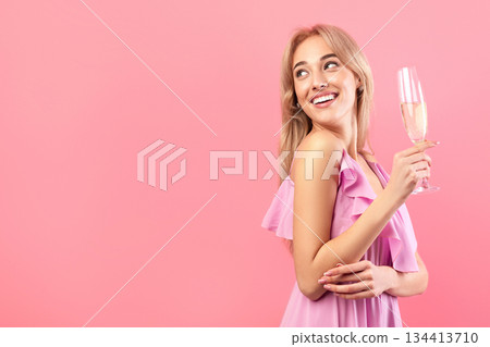 Beautiful blonde woman in dress holding glass of sparkly champagne, celebrating holiday, looking aside at empty space over pink studio background. Festive concept Beautiful blonde woman in dress holding glass of sparkly champagne, celebrating holiday, looking aside at empty space over pink studio background. Festive concept 134413710