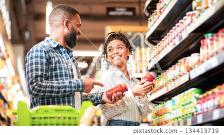 Happy African Family Couple Buying Food In Supermarket, Choosing Products Walking With Cart Along Aisles And Full Shelves Purchasing Groceries Together. Black Spouses Purchasing Essentials Together Happy African Family Couple Buying Food In Supermarket, Choosing Products Walking With Cart Along Aisles And Full Shelves Purchasing Groceries Together. Black Spouses Purchasing Essentials Together 134413719