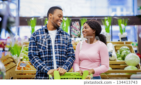 Portrait of smiling young black couple with shopping cart looking at each other in veggies section of grocery store. Affectionate African American family buying fresh food together at mall Portrait of smiling young black couple with shopping cart looking at each other in veggies section of grocery store. Affectionate African American family buying fresh food together at mall 134413743
