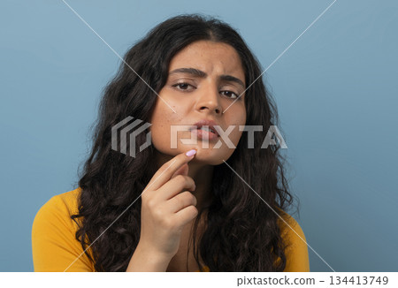 Stressed sad brunette young indian woman touching her face over blue studio background, having skin problems, closeup. Acne, pimples, wrinkles, dull skin 134413749