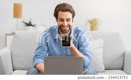 Cheerful Guy Using Voice Assistant On Mobile Phone Or Talking Using Loudspeaker Mode On Smartphone Sitting At Laptop Computer On Sofa At Home. Voice Search Application, Gadgets Lifestyle. 134413751