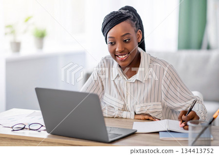 Young black lady having online training from home, using laptop and taking notes, copy space. Smiling african american woman attending online conference, writing tasks, living room interior Young black lady having online training from home, using laptop and taking notes, copy space. Smiling african american woman attending online conference, writing tasks, living room interior 134413752