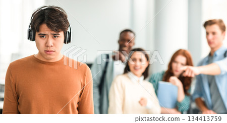 Abuse And Racism. Diverse People Bullying And Whispering Behind Back Of Unhappy Asian Man In Headset Standing Alone In Classroom, Pointing Fingers At Outcast Person, Fake Bad Friends. Selective Focus 134413759