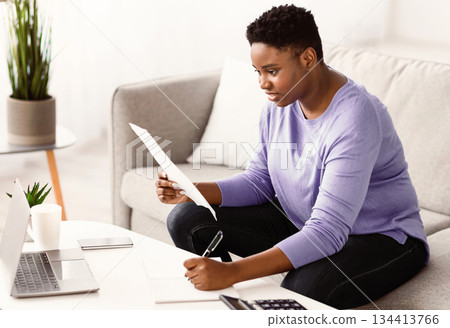 Doing Paperwork. Portrait of focused plus size young black lady working remotely at home office, holding paper and writing notes on financial report, sitting on the sofa at table with laptop 134413766