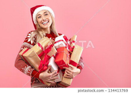 Smiling Caucasian woman in comfy Xmas sweater and Santa hat holding pile of gifts over pink studio background. Charming millennial lady with heap of presents celebrating winter holidays 134413769