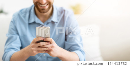 Happy Bearded Guy Using Smartphone Texting And Using Mobile Application On Phone Sitting On Couch At Home. Great App, Man Using Cellphone Browsing Internet Or Playing Game Indoor. Shallow Depth 134413782