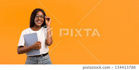 Cute young black lady touching her glasses, holding books or notebook, panorama with copy space, orange studio background. Education, studying, training, educational course concept 134413786