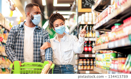 African American Couple In Protective Masks In Supermarket Doing Grocery Shopping, Buying Food Essentials Walking With Shop Cart In Groceries Store Indoors. Family Choosing Food Products On Shelves 134413789