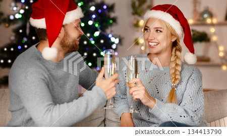 Happy man and woman in Santa hats celebrating New Year at home, drinking champagne Happy man and woman in Santa hats celebrating New Year at home, drinking champagne 134413790