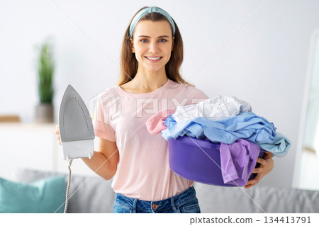 Joyful millennial housewife holding clean clothes and iron, ready to do domestic duties, indoors. Portrait of young woman prepared for ironing laundry, looking at camera and smiling Joyful millennial housewife holding clean clothes and iron, ready to do domestic duties, indoors. Portrait of young woman prepared for ironing laundry, looking at camera and smiling 134413791