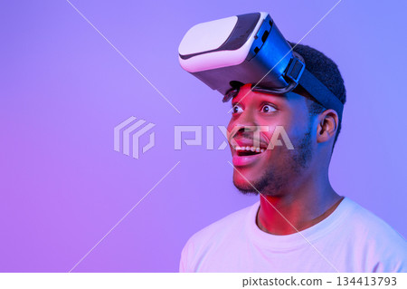 Video Gaming Concept. Portrait Of Amazed Funny Black Man Wearing VR Glasses Looking Away Over Purple Background, Excited African American Male Enjoying Virtual Reality Experience Under Neon Lighting 134413793