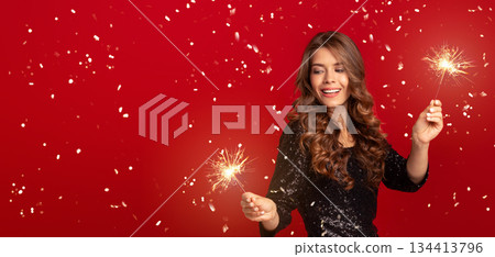 Beautiful millennial woman in elegant dress celebrating New Year with sparklers over red background 134413796