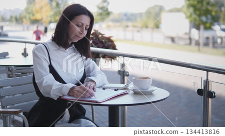 A business woman sits at a cafe table, writing notes and typing on a calculator while sipping coffee. She appears focused and engaged in her tasks under bright sunlight. A business woman sits at a cafe table, writing notes and typing on a calculator while sipping coffee. She appears focused and engaged in her tasks under bright sunlight. 134413816