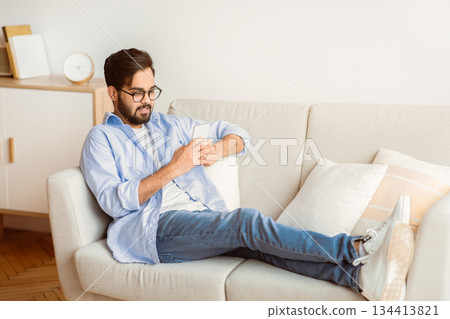 Domestic Leisure. Millennial Arab Guy Using Smartphone While Resting On Couch In Living Room, Smiling Middle Eastern Man Messaging On Cellphone While Sitting On Comfortable Sofa At Home, Free Space 134413821