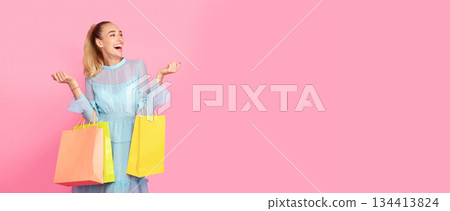 Big Sales Concept. Excited Woman In Dress Standing At Studio And Looking At Copy Space, Holding Bright Colorful Shopping Paper Bags, Laughing With Joy Isolated Over Pastel Pink Background Wall, Banner 134413824