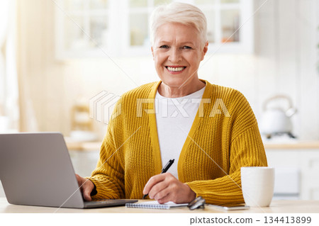 Online education, courses, webinar concept. Cheerful elderly woman sitting in front of laptop in kitchen, taking notes and smiling, drinking tea, enjoying studying online from home, copy space 134413899