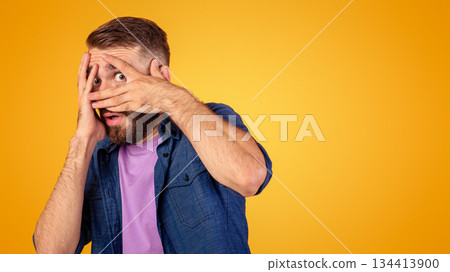 Scared Caucasian guy hiding behind his hands, peeping between fingers on orange studio background. Horrified young man suffering from phobia, feeling spooked. Negative emotions concept Scared Caucasian guy hiding behind his hands, peeping between fingers on orange studio background. Horrified young man suffering from phobia, feeling spooked. Negative emotions concept 134413900