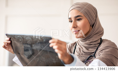 Portrait of muslim arab female medical doctor looking at xray scan, clinic interior, copy space. Young woman in hijab physician holding CT scan, checking disease progress or treatment efficiency Portrait of muslim arab female medical doctor looking at xray scan, clinic interior, copy space. Young woman in hijab physician holding CT scan, checking disease progress or treatment efficiency 134413906