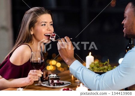 Loving African American Man Feeding His White Girlfriend With Cake On Romantic Date In Restaurant, Happy Interracial Lovers Celebrating Valentine's Day Together, Drinking Wine And Eating Dessert Loving African American Man Feeding His White Girlfriend With Cake On Romantic Date In Restaurant, Happy Interracial Lovers Celebrating Valentine's Day Together, Drinking Wine And Eating Dessert 134413908