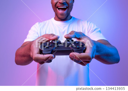 Excited african american guy playing video games with joystick, joyful black man enjoying modern entertainment, having fun in vivid neon light on purple studio background, selective focus, copy space Excited african american guy playing video games with joystick, joyful black man enjoying modern entertainment, having fun in vivid neon light on purple studio background, selective focus, copy space 134413935