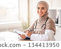 Female doctor taking anamnesis while having conversation with patient. Smiling muslim woman doctor in headscarf taking notes in medical chart, sitting at workdesk at her cabinet in clinic, copy space 134413950