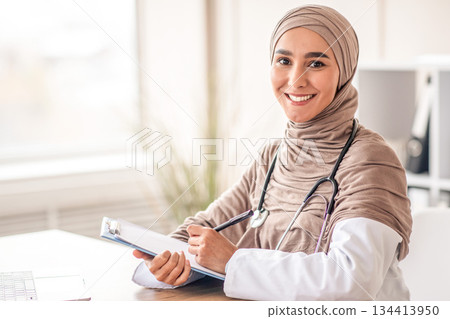 Female doctor taking anamnesis while having conversation with patient. Smiling muslim woman doctor in headscarf taking notes in medical chart, sitting at workdesk at her cabinet in clinic, copy space 134413950