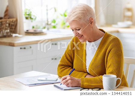 Positive elderly lady freelancer working from home or studying, sitting in kitchen with laptop and smartphone, taking notes while reading financial report, smiling at camera, copy space Positive elderly lady freelancer working from home or studying, sitting in kitchen with laptop and smartphone, taking notes while reading financial report, smiling at camera, copy space 134414051