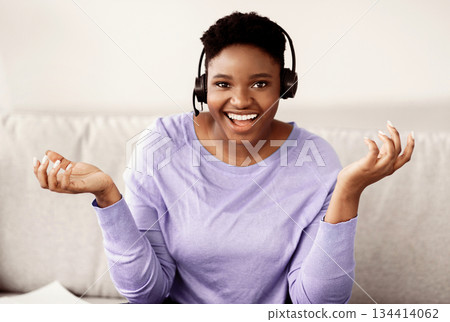 Video Conference. Front portrait of smiling young black woman having web call or virtual meeting, talking to camera while sitting at home office, wearing wireless headset, explaining Video Conference. Front portrait of smiling young black woman having web call or virtual meeting, talking to camera while sitting at home office, wearing wireless headset, explaining 134414062