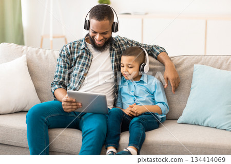 Young Black Father And Little Son Using Tablet Computer Together Wearing Wireless Headphones Watching Movie Sitting On Couch At Home. Family Of Two Browsing Internet Using Gadget Concept Young Black Father And Little Son Using Tablet Computer Together Wearing Wireless Headphones Watching Movie Sitting On Couch At Home. Family Of Two Browsing Internet Using Gadget Concept 134414069