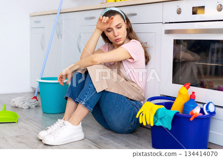 Depressed young housewife feeling tired from domestic chores, sitting on floor at kitchen. Young lady with cleaning supplies exhausted from house cleaning, suffering from OCD disorder Depressed young housewife feeling tired from domestic chores, sitting on floor at kitchen. Young lady with cleaning supplies exhausted from house cleaning, suffering from OCD disorder 134414070