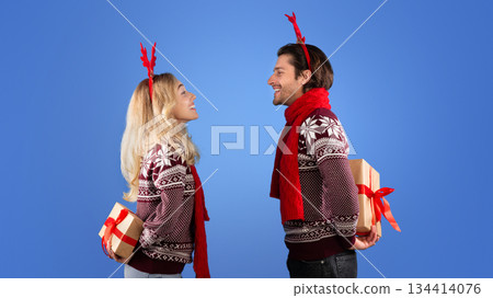 Side view of joyful couple hiding Christmas gifts behind their backs on blue background Side view of joyful couple hiding Christmas gifts behind their backs on blue background 134414076
