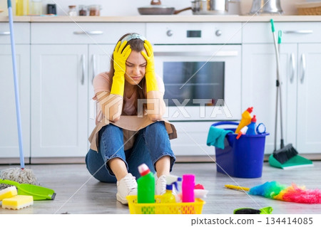 Desperate young housewife tired from domestic duties grabbing her head on floor at kitchen, full length portrait. Millennial woman feeling exhausted from housework. OCD concept Desperate young housewife tired from domestic duties grabbing her head on floor at kitchen, full length portrait. Millennial woman feeling exhausted from housework. OCD concept 134414085