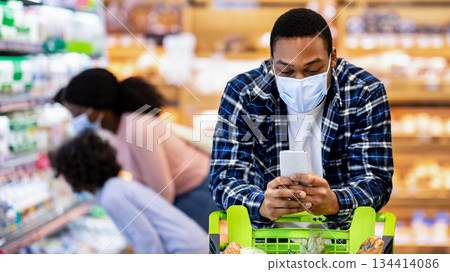 Cool black guy in face mask checking grocery list on smartphone while his wife and daughter choosing products at dairy department og huge modern supermarket, copy space Cool black guy in face mask checking grocery list on smartphone while his wife and daughter choosing products at dairy department og huge modern supermarket, copy space 134414086