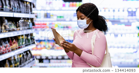 Millennial black lady with tote bag wearing face mask, shopping for groceries at mall. Young African American woman making choice of products, purchasing food at supermarket during covid outbreak Millennial black lady with tote bag wearing face mask, shopping for groceries at mall. Young African American woman making choice of products, purchasing food at supermarket during covid outbreak 134414093