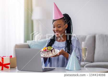 Positive african american woman in party hat showing her online friend cake, celebrating birthday online during self-isolation, happy black lady having video conference with family or boyfriend Positive african american woman in party hat showing her online friend cake, celebrating birthday online during self-isolation, happy black lady having video conference with family or boyfriend 134414098